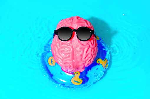 Cute character, made from a human brain model wearing sunglasses, enjoys a carefree summer moment as it floats on an inflatable tube in the water. Creative leisure and mental well-being concept.