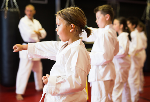 Group of girls and boys in kimono doing kata with their trainer in gym.