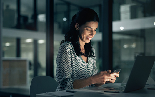 Night, woman working and phone with a smile in a office with planning and digital research. Startup, working and laptop with female person in a company with text reading and mobile networking in dark