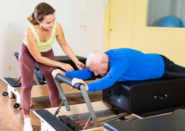 female trainer controlling senior man doing pilates on reformer - senior adult physical therapy gymnastics cheerful imagens e fotografias de stock