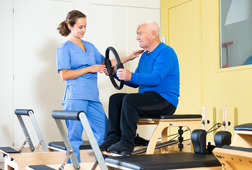 Professional female physiatrist controlling older man exercising with pilates ring to strengthen muscles. Physical medicine and rehabilitation concept