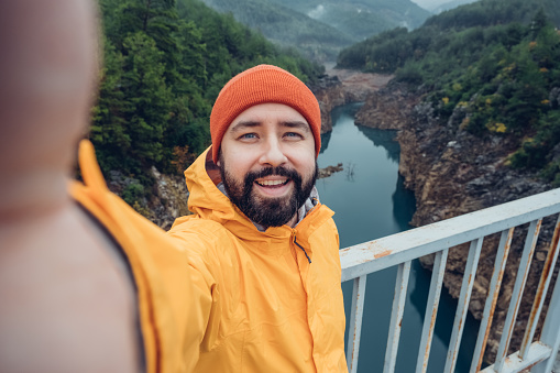 Hiker man taking selfie portrait on top of mountain. Guy in red hat and yellow jacket making video report or shooting video content for his travel vlog