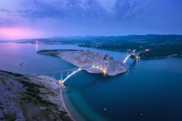 puente sobre el mar al atardecer. vista aérea del moderno puente krk con iluminación, isla, montañas, mar azul, barco y cielo púrpura por la noche. vista superior del dron de la carretera, las luces de la ciudad, las rocas. arquitectura - morning croatia blue sea fotografías e imágenes de stock