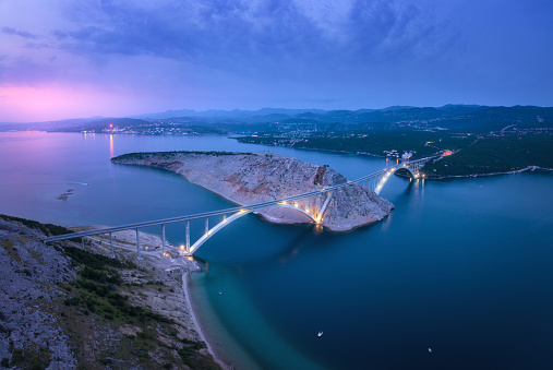 Bridge over the sea at sunset. Aerial view of modern Krk bridge with illumination, island, mountains, blue sea, boat and purple sky at night. Top drone view of road, city lights, rocks. Architecture
