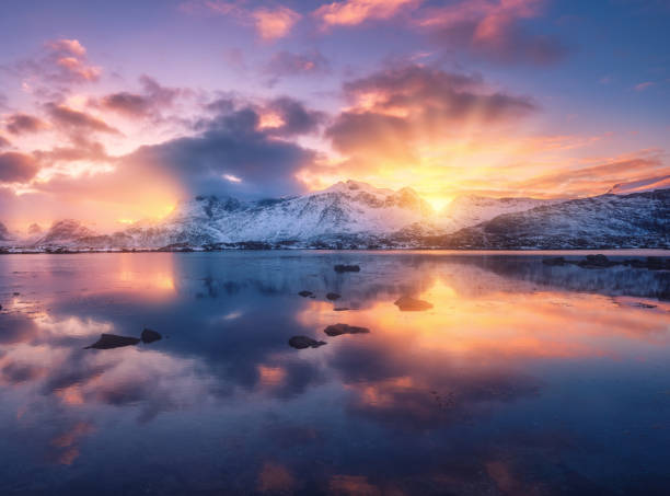 Sea coast, beautiful snowy mountains and colorful sky with clouds and golden sunlight at sunset in winter. Lofoten islands, Norway. Landscape, rocks in snow, reflection in water at dusk. Scenery stock photo
