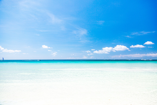 A seascape at the White Sand Beach on Boracay Island in the Philippines. The image captures the view of the sea from the beach, where the bottom of the frame displays shallow, transparent water. Moving towards the horizon, the water gradually transitions from turquoise to dark blue. Small ripples can be seen on the surface of the water. In the distance, white boats are visible on the horizon. The sky above is blue with scattered clouds. This photograph offers a glimpse of the natural beauty of Boracay Island's White Sand Beach and the serene coastal atmosphere it provides.