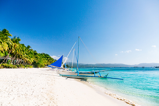 Boracay Island's famous white sand beach on a sunny day. The beach features clean, white sand stretching across the frame. Numerous palm trees can be seen along the shoreline. The sea appears calm and displays crystal-clear waters with a color palette transitioning from turquoise near the shore to dark blue in the distance. The sky is clear, revealing a vibrant blue hue without any clouds. Positioned at the center of the image are white boats parked on the beach, adding a visual element to the overall scene.