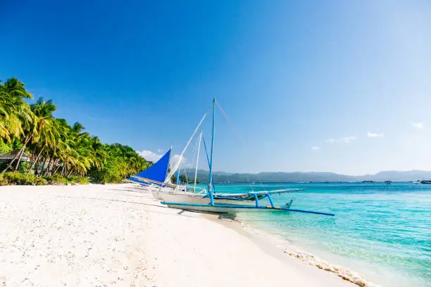 Boracay Island's famous white sand beach on a sunny day. The beach features clean, white sand stretching across the frame. Numerous palm trees can be seen along the shoreline. The sea appears calm and displays crystal-clear waters with a color palette transitioning from turquoise near the shore to dark blue in the distance. The sky is clear, revealing a vibrant blue hue without any clouds. Positioned at the center of the image are white boats parked on the beach, adding a visual element to the overall scene.