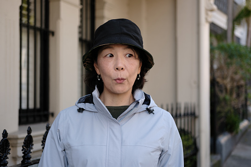 Asian woman with hat and jacket walking along a suburban street with a confused expression.