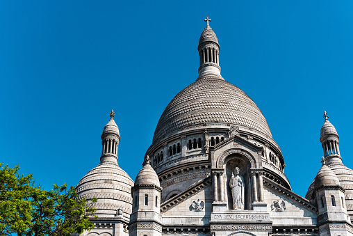 View of the Sacré-Coeur Basilica de Montmartre with clear blue sky - Paris, France. The Sacré-Cœur Basilica de Montmartre (Sacred Heart of Montmartre) is a Roman Catholic church and minor basilica in Paris dedicated to the Sacred Heart of Jesus. It was formally approved as a national historic monument by the National Commission of Patrimony and Architecture on December 8, 2022. It is located at the summit of the butte of Montmartre. From its dome two hundred meters above the Seine, the basilica overlooks the entire city of Paris and its suburbs. It is the second most popular tourist destination in the capital after the Eiffel Tower.\nThe porch of the south façade is topped with a statue representing the Sacred Heart of Christ.