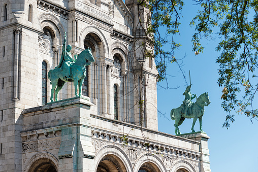 Facade of the Sacré-Coeur Basilica de Montmartre and view of King Saint Louis IX and Saint Joan of Arc equestrian statues. The Sacré-Cœur Basilica de Montmartre (Sacred Heart of Montmartre) is a Roman Catholic church and minor basilica in Paris dedicated to the Sacred Heart of Jesus. It is located at the summit of the butte of Montmartre in Paris, France. It is the second most popular tourist destination in the capital after the Eiffel Tower.\nThe arches of the façade are decorated with two equestrian statues of French national saints Joan of Arc and King Saint Louis IX, both executed in bronze by Hippolyte Lefèbvre.