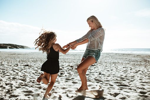Happy woman playing with a girl on the beach sand. Mother and daughter holding hands and having fun together on the seashore.