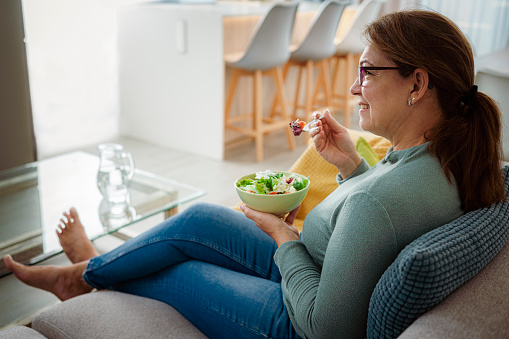 Mature hispanic woman eating healthy salad sitting on sofa