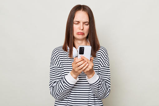 Sad upset brown haired adult woman wearing striped shirt standing isolated over gray background using phone with unhappy expression reading message with sorrow.