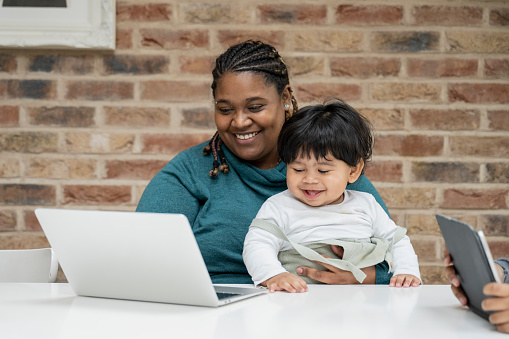 Multiracial businesswoman in early 30s, casually dressed, sitting at dining table with 15 month old baby in her lap and using laptop.