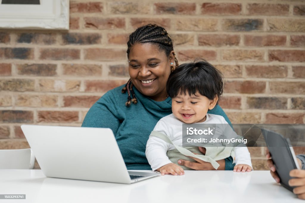 Cheerful mother multi-tasking at home Multiracial businesswoman in early 30s, casually dressed, sitting at dining table with 15 month old baby in her lap and using laptop. Child Stock Photo