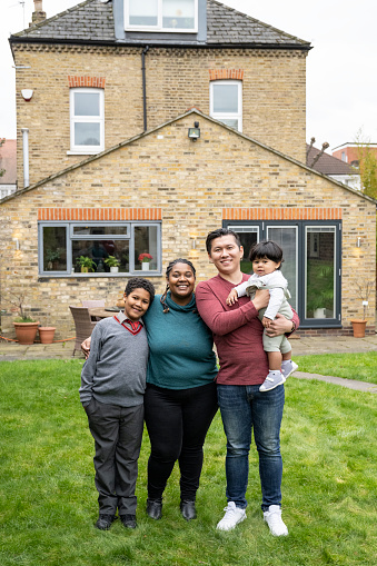 Full length front view of mid adult couple standing in back garden with 10 year old schoolboy in uniform, 15 month old in arms, all smiling at camera.