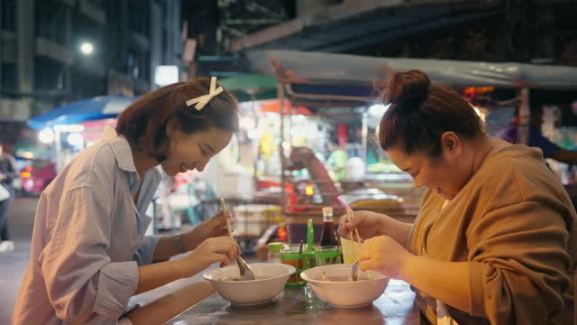 Two Asian women enjoy eating street noodles at a night market.