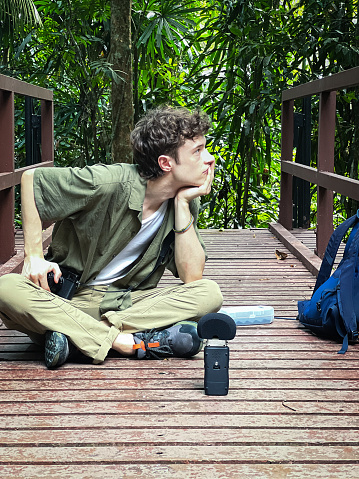 A young man biologist sitting on a wooden bridge with a camera and sound recorder to capture bird and insect sounds in a jungle area located in a public park.  Penang, Malaysia.