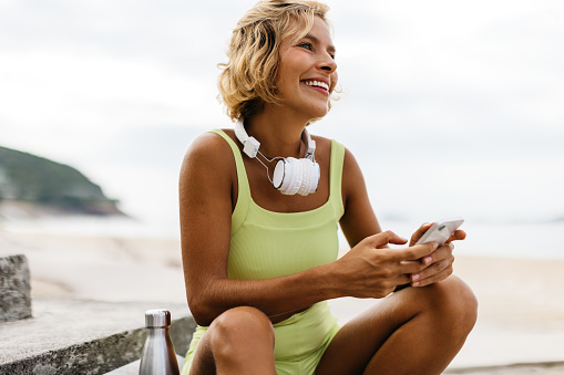 Caucasian woman in workout clothing takes a break from her active routine outdoors, using her phone to browse the internet. Happy young woman using technology to navigate her fitness journey.