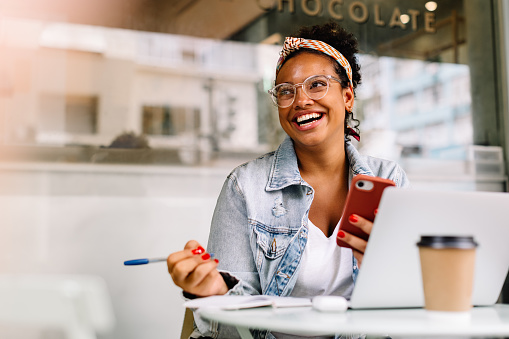 Happy post-graduate student working on her assignment at a café, browsing with her phone. Young woman enjoying the quiet atmosphere in a coffee shop as she engages in her school work.