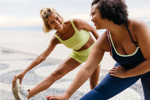 Two women with different body sizes doing stretch exercises during a beach workout session. Happy female friends warming up for a jog along along the ocean promenade in fitness clothing.