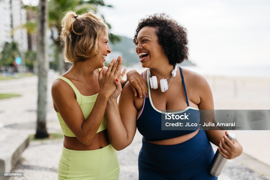 Fitness women laughing and high-fiving after a beach workout Two happy women in sportswear enjoy their workout routine on the ocean promenade, celebrating with a high-five. Female friends with different body shapes embracing a lifestyle of fitness. Women Stock Photo