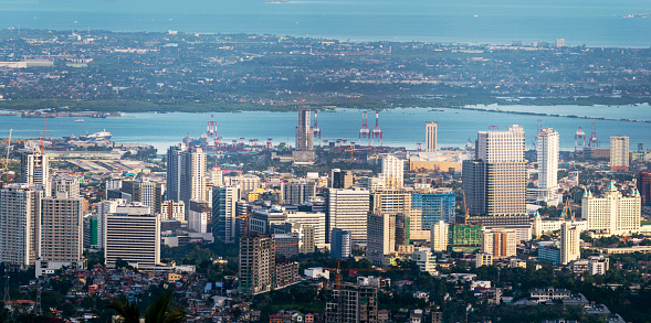 Pre-sunset sunlight bathes the taller buildings of Cebu's capital ,looking across the sea channel to Maktan Island,over palm trees and lush foliage,from the mountainside viewing point of the temple.