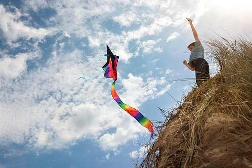 Boy flying a kite on beach sand dune in a windy blue sky background