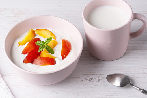 Fresh cottage cheese in pink bowl with mint leaves and peach pieces and milk on white background.