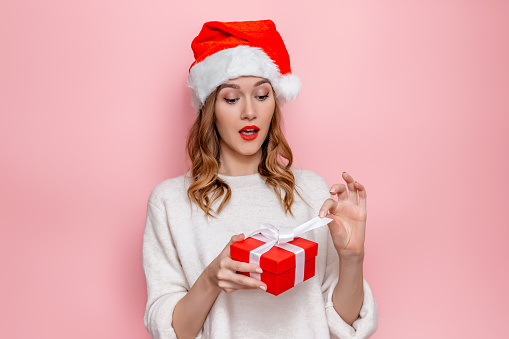 Funny surprised shocked young woman in santa hat opens red gift box and holds ribbon in her hands isolated on pink studio background