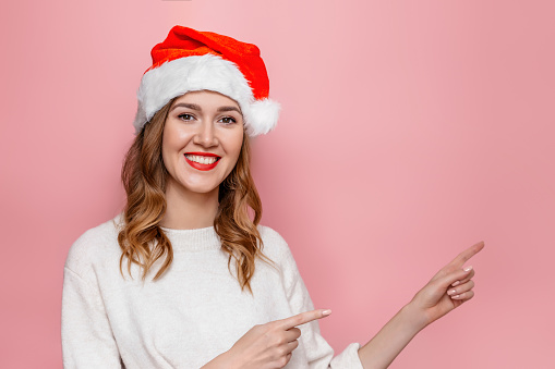 Young happy caucasian woman in santa hat smiling and pointing finger at copy space isolated on pink studio background