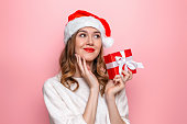 New year gifts concept. Cute caucasian young girl in a santa hat holds a box with a gift thinks and dreams makes a wish isolated on a pink background in the studio.