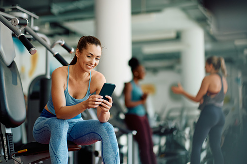Happy athletic woman texting on cell phone while exercising in a gym. Copy space.