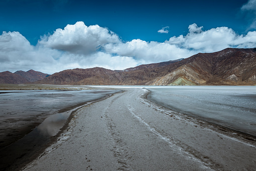 A walkway on the fully frozen Pangong Lake, where a sunlit day illuminates the pristine winter scenery. Surrounded by a vast blue sky and gentle white clouds, immerse yourself in the serene ambiance of this breathtaking landscape