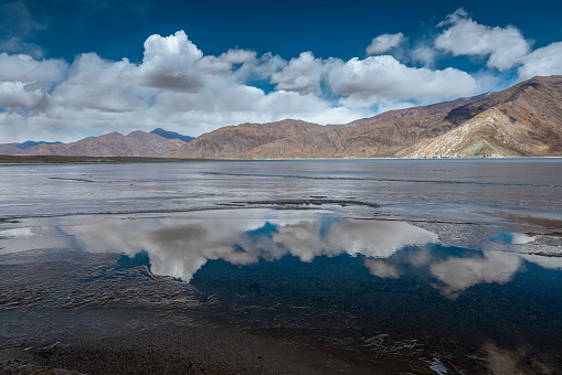 A stunning winter landscape of Pangong Lake showcasing the magical reflection of clouds on its mostly frozen surface. As the lake begins to thaw, a mesmerizing play of partially melted water unveils its icy charm under a sunny blue sky adorned with fluffy white clouds