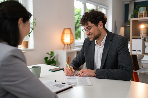 Young Caucasian man, signing the contract during meeting with a female real estate agent of Japanese ethnicity at the office