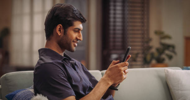 Indian Man Using Smartphone at Home: Seamlessly Managing Personal Finances, Exploring New Recipes, Staying Updated with the Latest News, and Embracing the Versatility of Modern Technology. Medium Shot