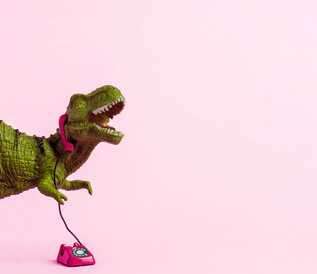 Cute green toy dinosaur speaking on retro phone on pink background. Copy space.