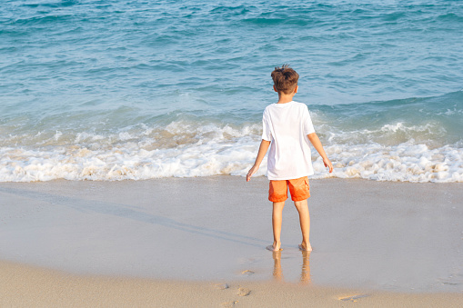 Child standing on the sandy beach, boy looks into the blue sea, concept of summer vacation and relaxation.