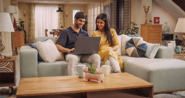 Portrait of Loving Young Indian Couple Enjoys Browsing Online Shopping Websites Together On A Laptop, While Sitting Cozily On Their Living Room Sofa. Browsing Interner, Choosing Products to Purchase retrato de una joven pareja india amorosa disfruta navegando juntos por sitios web de compras en línea en una computadora portátil, mientras están sentados cómodamente en el sofá de su sala de estar. navegación interna, elección de productos para co