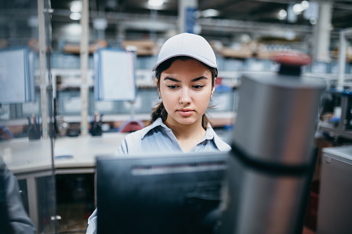 Experience the dedication and focus of a young female worker or engineer as she diligently works at her computer monitor in the factory. This compelling image captures her expertise and concentration as she handles important tasks and monitors crucial data. Witness the seamless integration of technology and skill as she navigates the complexities of the industrial environment.