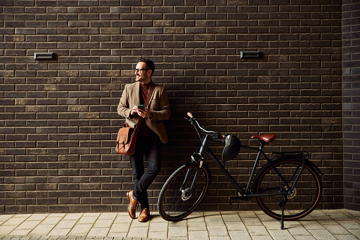 A smiling businessman with glasses on, holding a phone, and standing outside with a bicycle, looking around.