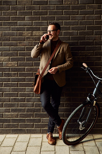 A cheerful businessman with a bicycle leaning on a brick wall and talking on a mobile phone.