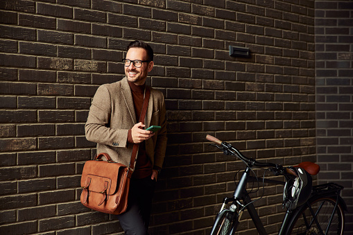 A happy businessman with glasses on standing by his bicycle in front of the brick wall and holding a mobile phone.
