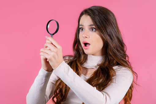 Portrait of a surprised pretty girl looking through magnifying glass on the pink background.