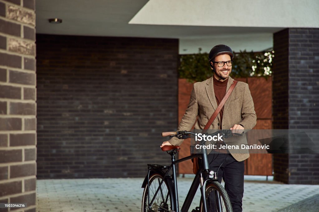 A happy businessman with a helmet, walking on foot with a bicycle, outdoors. A cheerful businessman in a suit walking on foot with a bicycle with a helmet on his head. Cycling Stock Photo