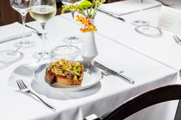 istock Buckwheat bread bruschetta with tomato paste, celery leaves, capers and artichokes. Bruschetta stands on a light ceramic plate on a white tablecloth. Nearby is a glass of white wine, a vase and several empty glasses. 1503346491