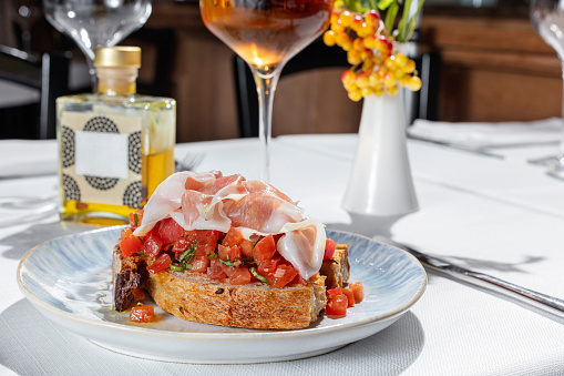 Buckwheat bread bruschetta with prosciutto and chopped tomatoes with herbs. Bruschetta stands on a light ceramic plate on a white tablecloth. Nearby is a glass of rose wine, a vase and several empty glasses.