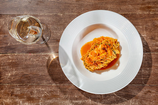 Spaghetti in tomato sauce with basil and shrimp. The food lies in a light ceramic plate, next to it is a glass of white wine. Dishes are placed on a wooden table.
